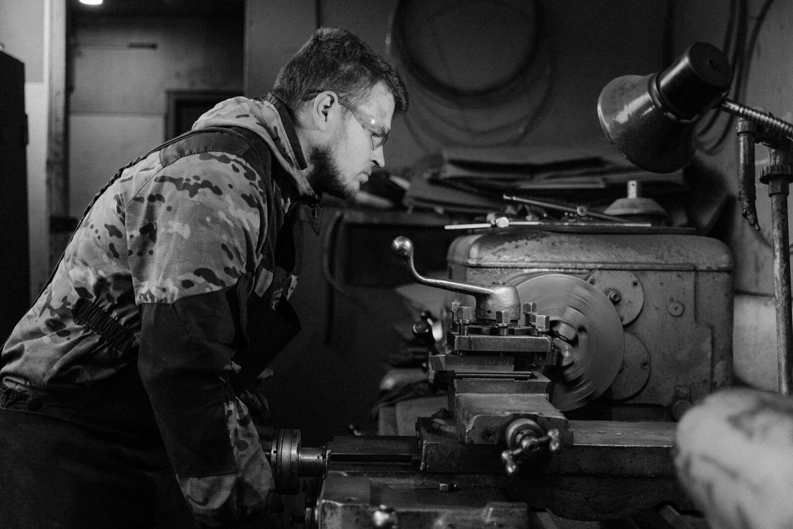 A bearded man concentrating on operating a lathe machine in a workshop, monochrome.